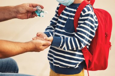 Koronavirüs pandemisi sırasında sırt çantalı küçük çocuğun eline plastik şişeden antiseptik döken yüksek açılı bir adam.