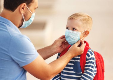 Tanınmayan baba, Coronavirus pandemi sırasında okula hazırlanırken neşeli okul çocuğunun yüzüne tıbbi maske takıyor.