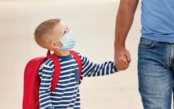Positive schoolboy in medical mask with backpack   holding hand of unrecognizable dad   while walking on stree