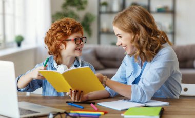 Genç bir anne, gözlüklü oğluna ödevlerinde yardım ediyor, oğlunun not defterine bakıyor, evde birlikte ders çalışırken.