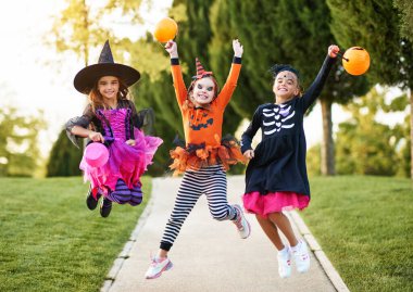 Full body delighted girls in spooky costumes smiling and leaping up on path during Halloween celebration in par clipart