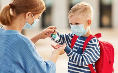 Korunaklı maskeli annenin Coronavirus pandemi sırasında küçük okul çocuğunun ellerine dezenfektan dökerken yan görüntüsü