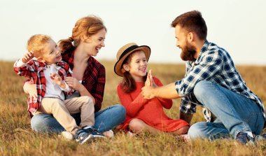 Mutlu aile: anne, baba, çocuk, oğul ve kız çimenlerde oturur, güler ve güneşin doğasında oynarlar.