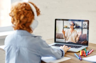 Kulaklıklı isimsiz bir çocuk masada oturuyor ve dost canlısı bir öğretmenin gülüşünü dinliyor ve evdeki online derste ilginç hikayeler anlatıyor.