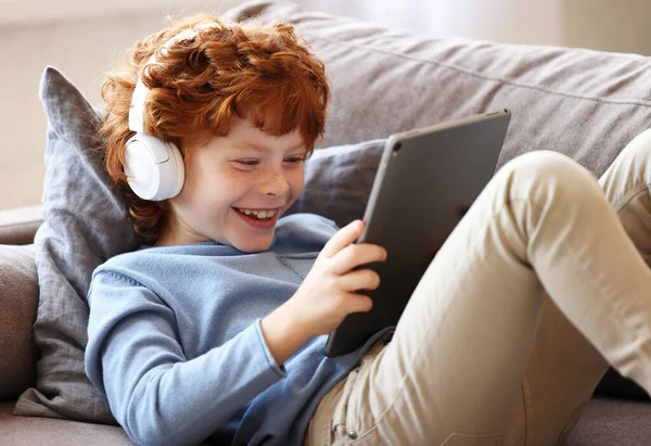坐在沙发上 头戴耳机 脚交叉坐在沙发上 用平板电脑在客厅里 快乐的小姜子笑了 — 图库照片