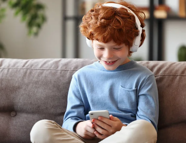 座っている間に笑顔のヘッドフォンで陽気な小さな生姜の男の子はソファに足を踏み 自宅でスマートフォンを使用して — ストック写真