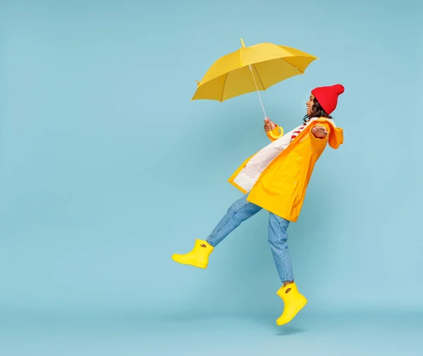 外衣姿の幸せな民族女性の側面図と傘の跳躍と青の背景に雨の秋の日に楽しみを持っている — ストック写真