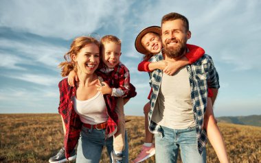 Mutlu aile: anne, baba, çocuk, oğul ve kız kucaklaşıyor, gülüyor ve çimenlerde eğleniyor, gülüyor ve mavi bir gökyüzüne karşı doğada oynuyor.