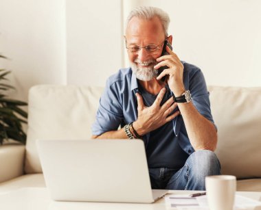 Kanepede oturup laptodan veri okurken akıllı telefondan gülümseyen ve konuşan mutlu sakallı kıdemli adam.