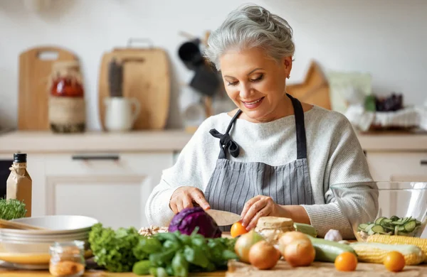 家庭のキッチンで新鮮な野菜と健康的な料理を準備しながら エプロン笑顔で陽気な中年主婦 — ストック写真