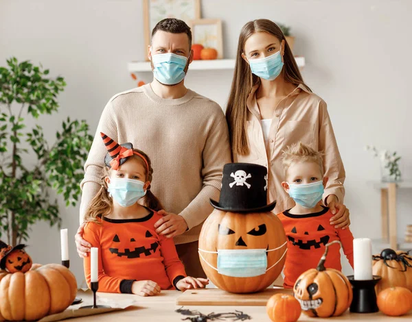 Cheerful family  in medical masks makes jack o lantern out of a pumpkin and  decorates house  in cozy kitchen during Halloween celebration at home during the covid19 coronavirus pandemi