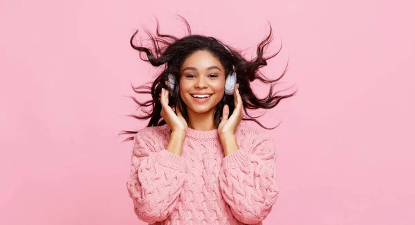 楽観的な若いですエスニック女性で編んだセーター笑顔と揺れ毛で音楽を聞きながらでヘッドフォンでピンクの背景 — ストック写真