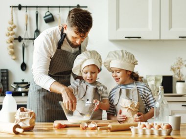 Mutlu küçük kardeşler aşçı şapkaları ve önlükleri içinde hamur işi için malzemeleri karıştırırken aynı zamanda evde gülen babayla yemek pişiriyorlar.