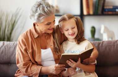 Mutlu yaşlı kadın ve küçük kız kanepede oturup evde büyüleyici peri masalları okurken gülümseyip birbirlerine bakıyorlar.
