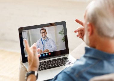 Yukarıdan yaşlı bir erkek doktorla video görüşmesi yapıyor ve evdeki kanepede otururken konuşuyor.