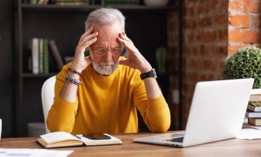 Şık kıyafetli, yorgun düşmüş, şakak okşayan ve evde rahat bir iş yerinde masada otururken düşünen bir adam.