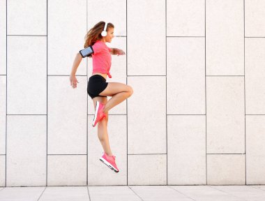 Spor giyim ve kulaklık takan genç ve enerjik bir bayanın tüm vücut görüntüsü ve telefon kol bandıyla duvara sıçrayan kulaklıklar.