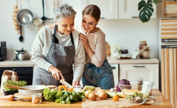 快乐快乐的家庭年轻女性拥抱成熟的母亲 同时在家里厨房里准备健康的菜和新鲜蔬菜 — 图库照片