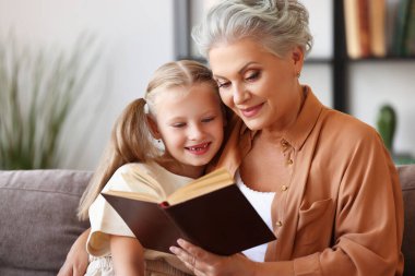 Mutlu yaşlı kadın ve küçük kız kanepede oturup evde büyüleyici peri masalları okurken gülümseyip birbirlerine bakıyorlar.