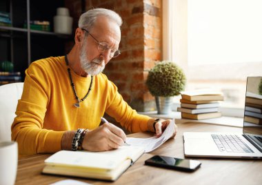 Gözlüklü ciddi bir kıdemli adam, evdeki rahat işyerinde masa başında evrak işleriyle uğraşırken belge üzerine notlar alıyor.