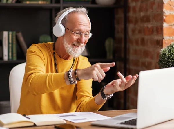 快乐的老年男性自由职业者 一边在耳机里一边说话一边弯着手指一边从家里打电话给同事 — 图库照片