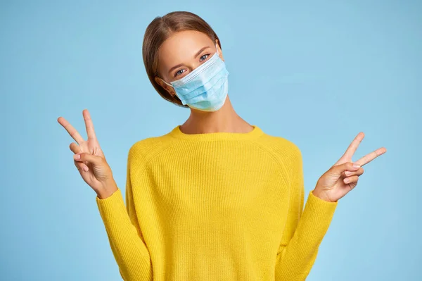 コロナウイルス パンデミの真ん中に青い背景をした保護医療マスクと黄色いセーターを着た美しい若い女性 — ストック写真
