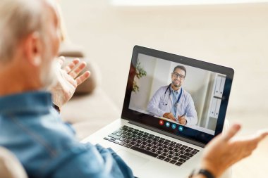 Yukarıdan yaşlı bir erkek doktorla video görüşmesi yapıyor ve evdeki kanepede otururken konuşuyor.