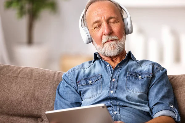头戴耳机 头戴平板电脑的老人坐在沙发上 闭着眼睛听音乐 在舒适的客厅里休息 — 图库照片