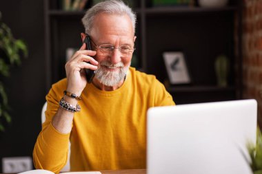 Evde rahat bir iş yerinde masa başında otururken gülümseyen ve telefona cevap veren şık yaşlı bir adam.