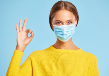 Korunaklı tıbbi maske ve sarı kazak giyen güzel genç bir kadın Coronavirus Pandemi 'nin ortasında mavi bir arka planda her şeyin iyi olacağını gösteriyor.