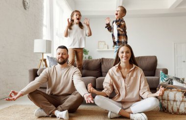 Mutlu aile: çocuklar evdeki kanepede zıplayıp eğlenirken huzurlu genç ebeveynler nilüfer pozisyonunda oturup meditasyon yapıyorlar.