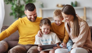 Mutlu aile genç ebeveynleri, günlük kıyafetleriyle gülümseyen çocuklarıyla kanepede oturup tablet okurken evde birlikte vakit geçiriyorlar.