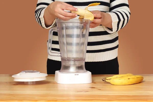 Young woman making  banana smoothie on gray background. She peels a banana  and mixing ingredients together  in blender. Simple elegant picture with copy space.