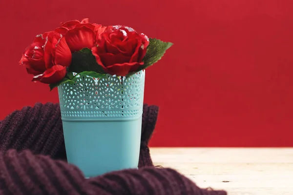 美丽的玫瑰花在塑料花瓶与紫色的冬天围巾在木桌子与红色背景 — 图库照片