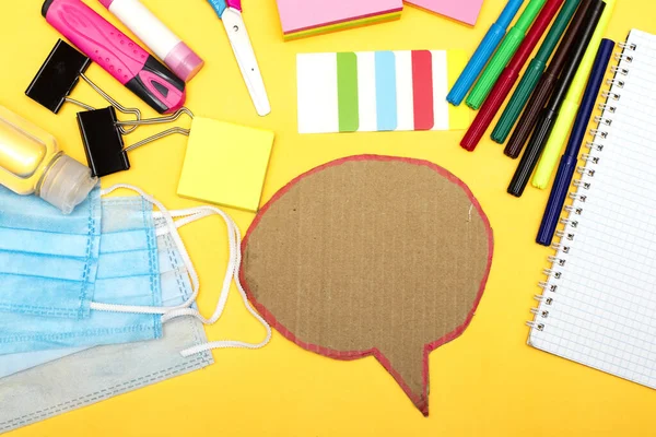 Back to school, school supplies with medical face mask and empty notebook and chat bubble on a yellow background. Protection of schoolchildren and students from the virus