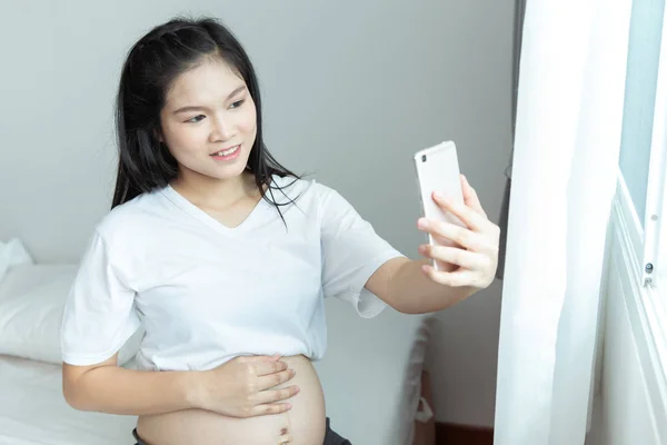 Veselá těhotná mladá Asianka seděla na posteli a vzala si s sebou — Stock fotografie