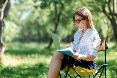Parkta bir katlanır sandalye üzerinde oturan ve bir defterde yazılı bir dizüstü bilgisayar ile genç kız
