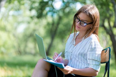 Parkta bir katlanır sandalye üzerinde oturan ve bir defterde yazılı bir dizüstü bilgisayar ile genç kız