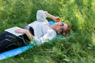 Kadın parkta çim üzerinde yalan ile bir dizüstü bilgisayar ve müzik dinleniyor