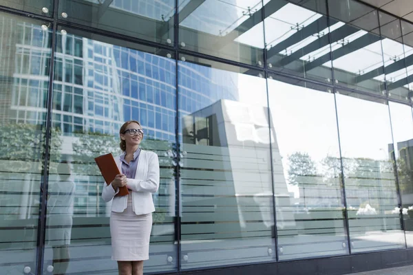 The young woman in gray office dress holds in hand folder with documents. Businesswoman standing next to the business center