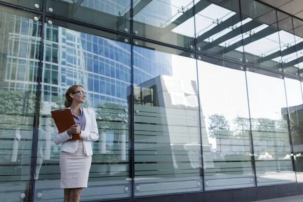 The young woman in gray office dress holds in hand folder with documents. Businesswoman standing next to the business center