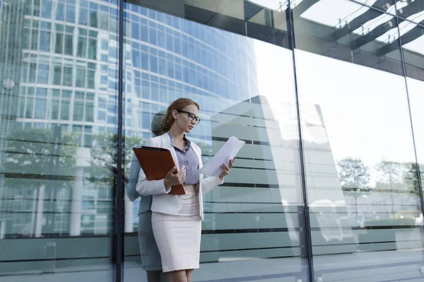 The young woman in gray office dress holds in hand folder with documents. Businesswoman standing next to the business center