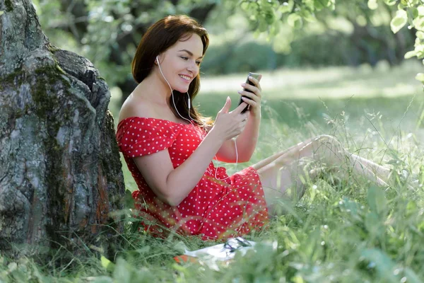 年轻漂亮的女人坐在树下的草地上 用她的智能手机 — 图库照片