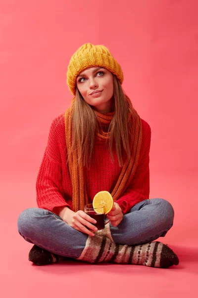 穿着红色毛衣和针织帽子的女孩拿着一杯热的酒和一层橙片。红色背景。演播室 — 图库照片