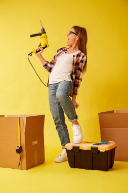 Taşıma kutuları arasında duran bir matkap tutan genç kadın. Stüdyo
