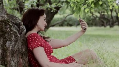 Artı boyutu kız ağaç altında çim oturan ve onu Smartphone kullanarak