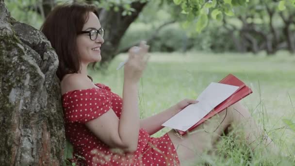 Niña sentada en la hierba bajo el árbol y escribiendo notas — Vídeo de stock