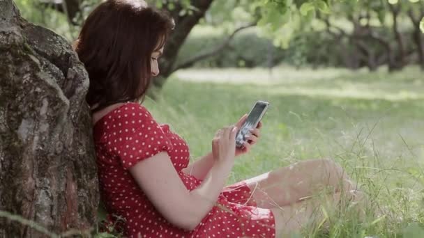 Plus size girl sitzt auf gras unter baum und benutzt ihr smartphone — Stockvideo