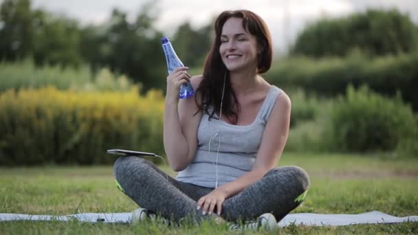 Fitness bela mulher bebendo água depois de se exercitar no parque na pose de lótus — Vídeo de Stock