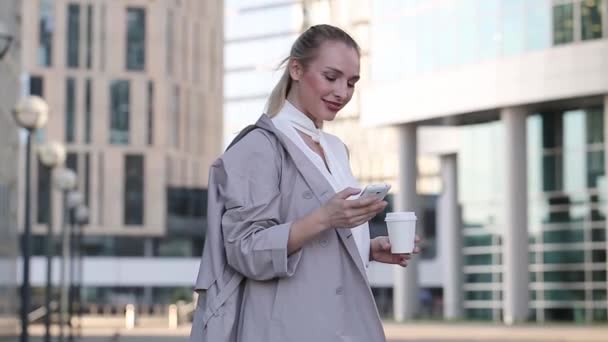 Joven mujer de negocios con taza de café usando su smartphone de pie entre el centro de la ciudad — Vídeos de Stock